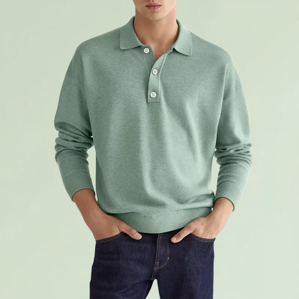 Person wearing a light green sweater over a collared shirt with dark jeans on a plain background