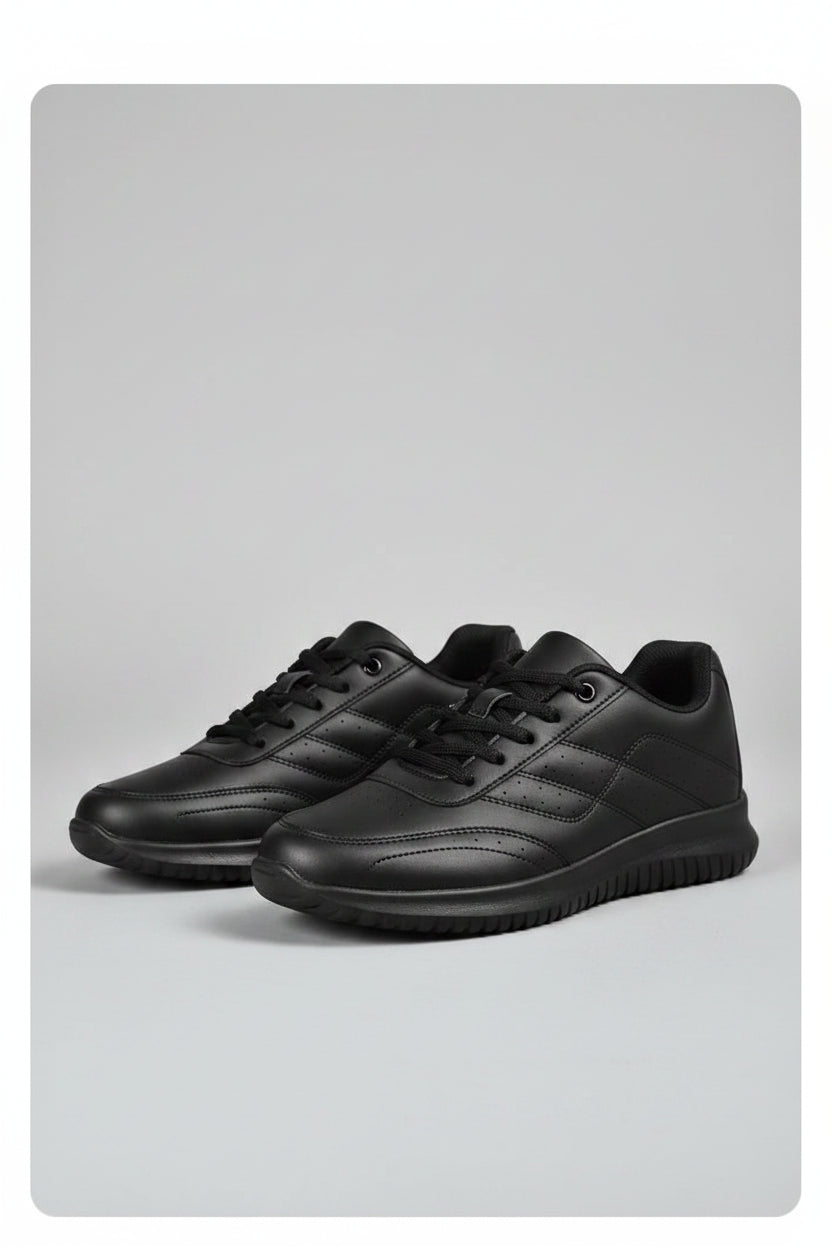 Black sneaker with a textured sole worn by a person in black pants on a concrete surface.