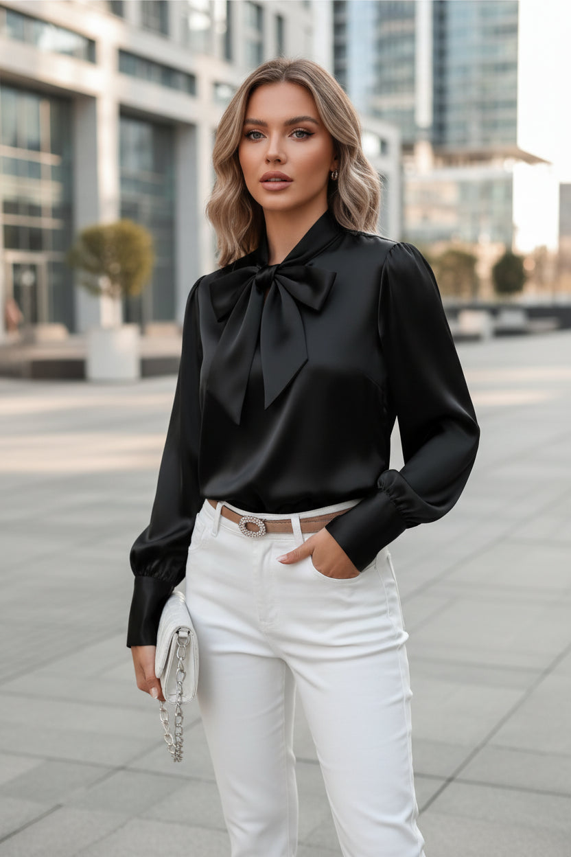 Black satin blouse with a large bow tie worn by a person, sitting on a white chair.