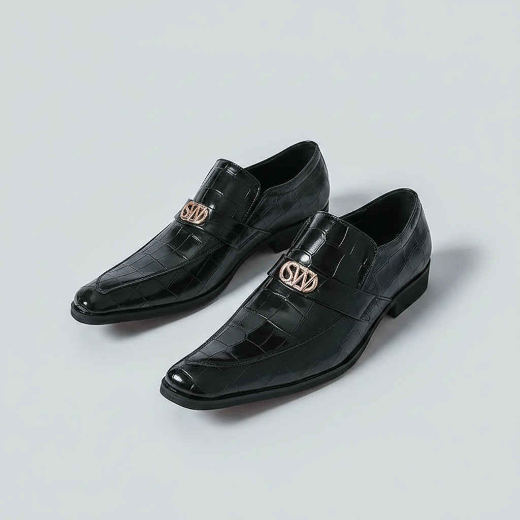 Black dress shoes with gold logo worn by a person on a white background