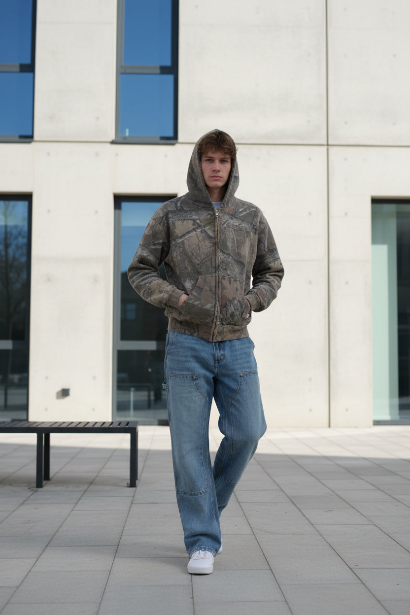 Person wearing a patterned shirt and jeans standing against a plain background