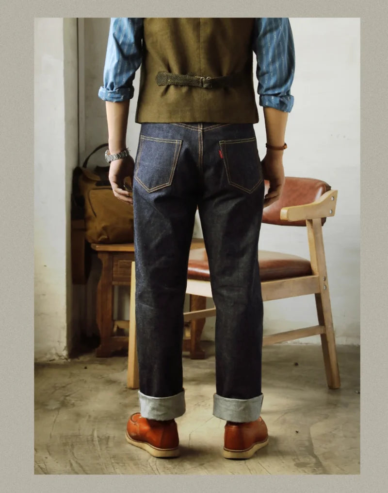 Person wearing a vest, blue shirt, dark jeans, and brown shoes in a room with a chair and bag.